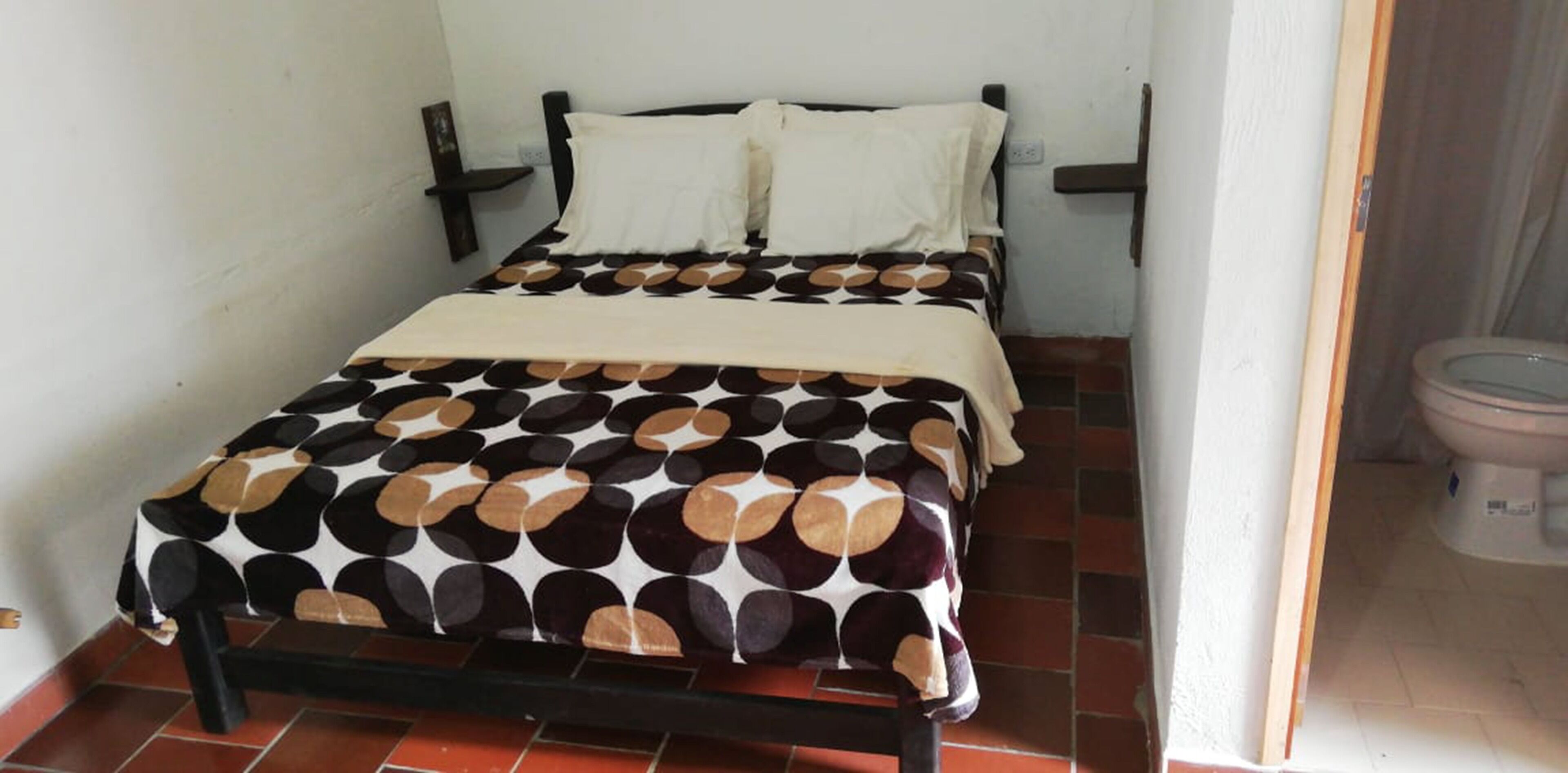 A small, single-bed room with a patterned bedspread, a bedside table, and a toilet visible through an open door.