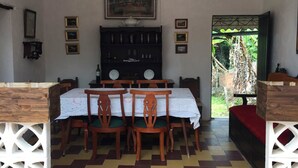 Uma sala de jantar com piso xadrez, cadeiras de madeira, uma mesa com toalha branca e um lustre.