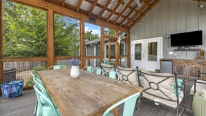Un área de comedor al aire libre, techada, con una mesa y sillas de madera, un televisor montado en la pared y vistas a los árboles circundantes.