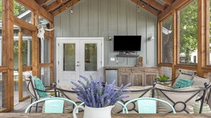 Un espacio de estar al aire libre techado, con televisor, vigas de madera y un ventanal grande.