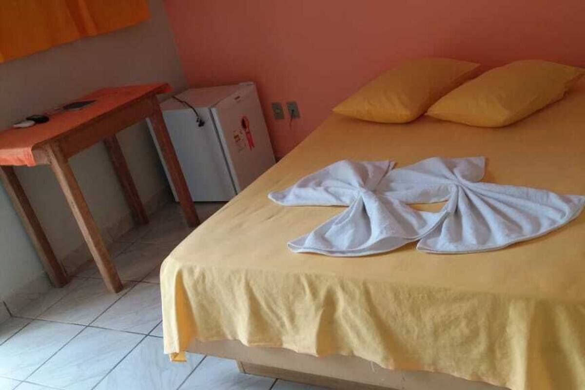 Una cama con cubrecama amarillo y dos almohadas, una mesa de madera con cubierta roja y un refrigerador blanco pequeño.