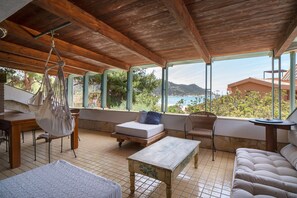 Een overdekt terras met uitzicht op zee, een houten tafel, een comfortabele bank en een hangstoel.
