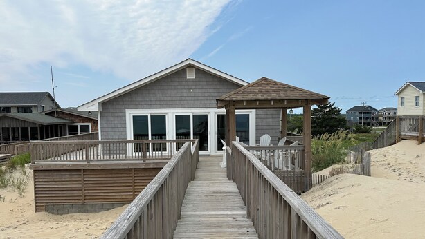 Una passerella in legno conduce a una casa sulla spiaggia con un portico coperto e un'ampia porta scorrevole in vetro.