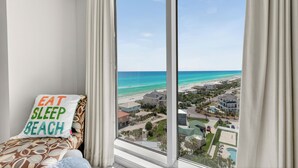 Un dormitorio con un ventanal que da a una playa, una almohada con la inscripción 'EAT SLEEP BEACH' y vistas a una zona turística.