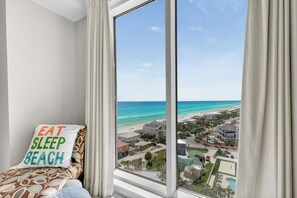 A bedroom with a large window overlooking a beach, a pillow with 'EAT SLEEP BEACH' text, and a view of a resort area.