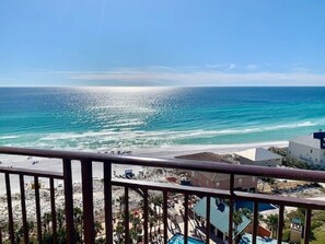 A beach view from a balcony with a railing, ocean, and buildings in the background.