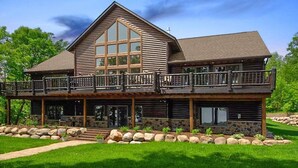 A large, two-story log cabin with a prominent balcony and stone foundation. - Luxurious Expansive Cabin Escape on Gull Lake! (Nisswa)