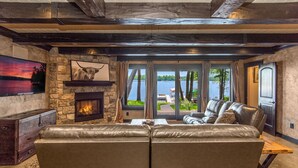 A cozy living room with a stone fireplace, leather sofa, and a large window overlooking a lake. - Luxurious Expansive Cabin Escape on Gull Lake! (Nisswa)