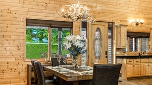 A wooden-paneled dining room with a chandelier, a table set for four, and a view of a green landscape through large windows. - Luxurious Expansive Cabin Escape on Gull Lake! (Nisswa)