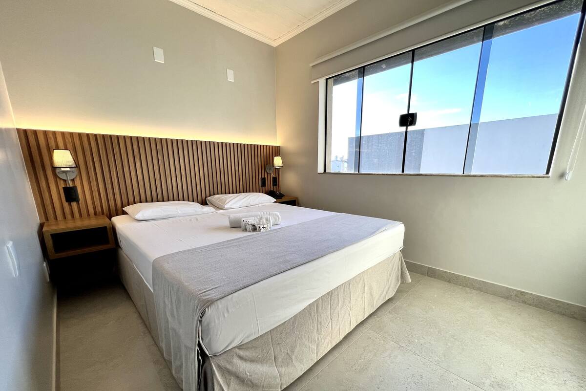 A hotel room with a large bed, a nightstand, a wall-mounted lamp, and a window with a view of the city.