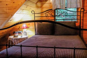 A bedroom with a sloped ceiling, a bed with a metal frame, a bedside table with a lamp, and a window with curtains.