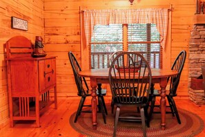A wooden cabin interior with a dining table, chairs, a wooden cabinet, and a window with a view of trees.