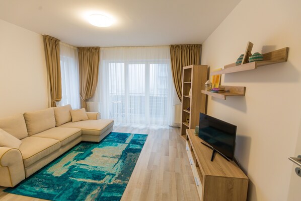 A modern living room with a beige sectional sofa, a flat-screen TV on a wooden stand, a blue patterned rug, and a wooden bookshelf.