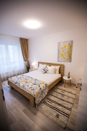 A bedroom with a wooden bed, white bedding, a floral-patterned bedspread, a small round table with a vase, and a painting on the wall.