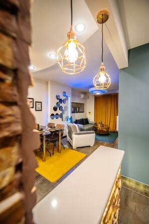 A modern living room with a dining area, featuring a white sofa, a coffee table, and a dining table with chairs. There are two pendant lights hanging from the ceiling.