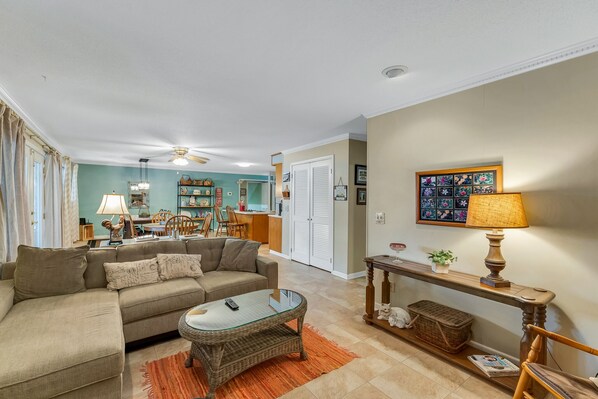 A living room with a sectional sofa, a glass-top coffee table, a wicker side table, and a lamp. There is a dining area in the background with a table and chairs. - Nanas Lakehouse - Private dock with Jet-ski port! (Jasper)