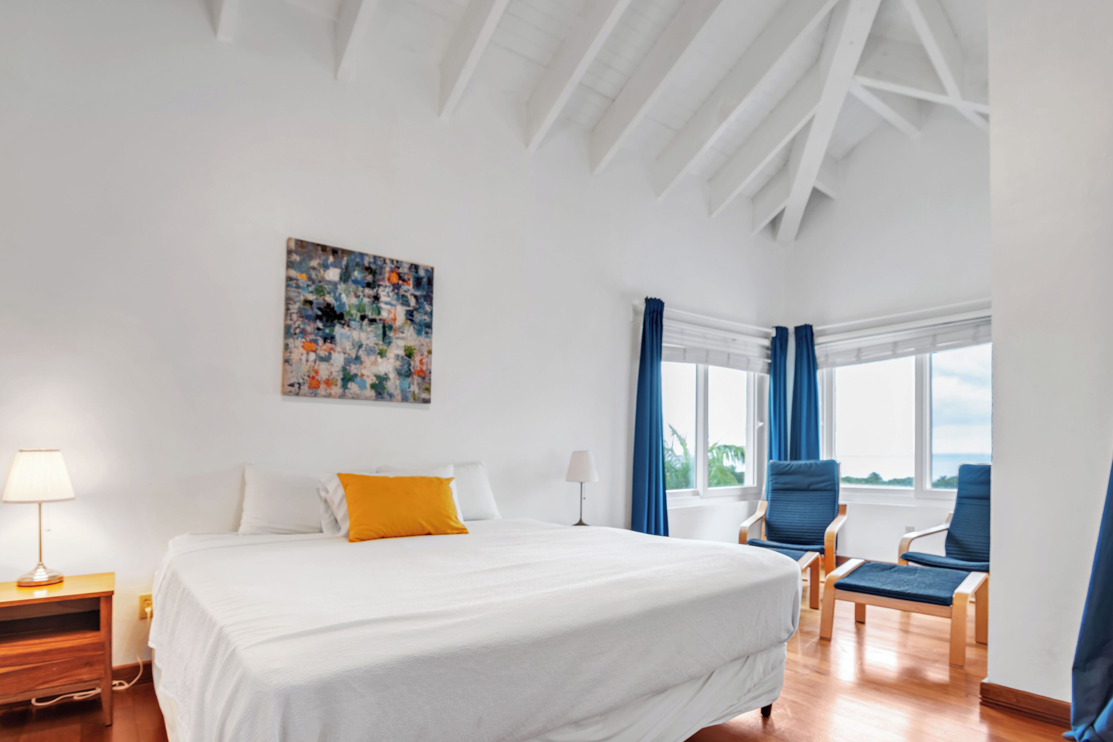 Quarto de hotel com uma cama grande, uma mesa com luminária, uma cadeira e uma janela com cortinas.