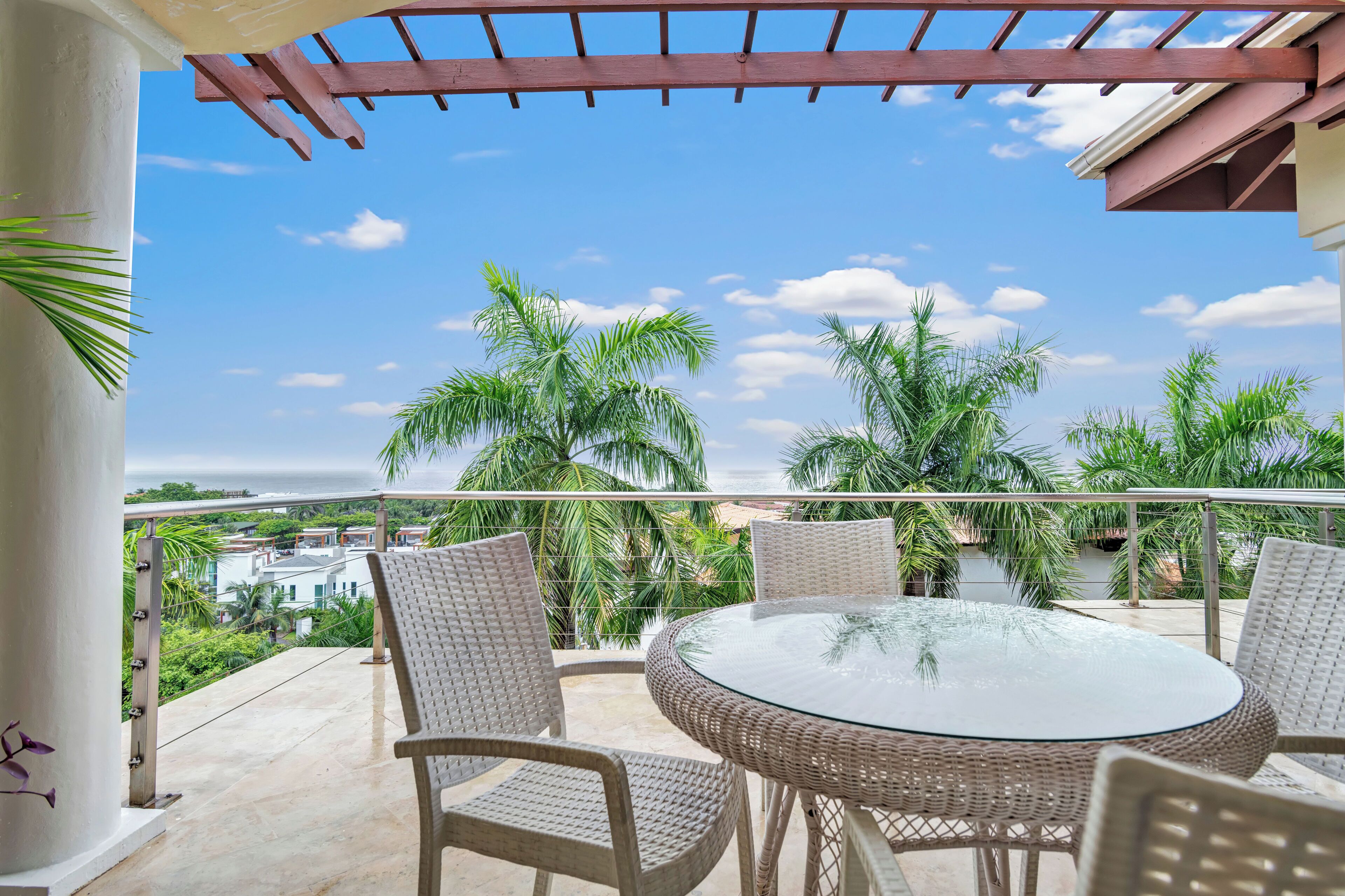 A balcony with wicker chairs and a glass table, overlooking a beach and palm trees.