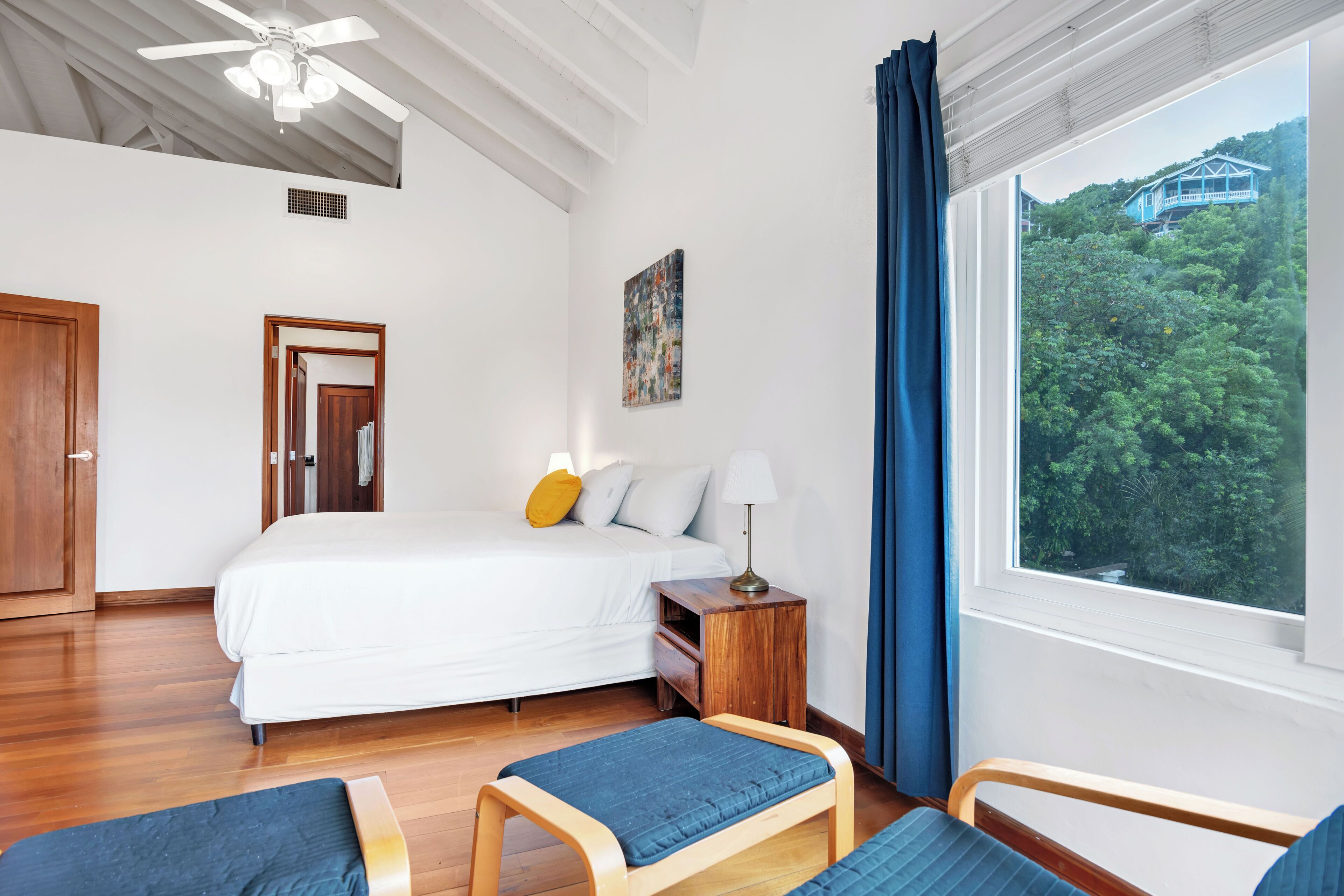 Ein Schlafzimmer mit Bett, Nachttisch, Lampe und Blick auf ein Haus durch ein Fenster.