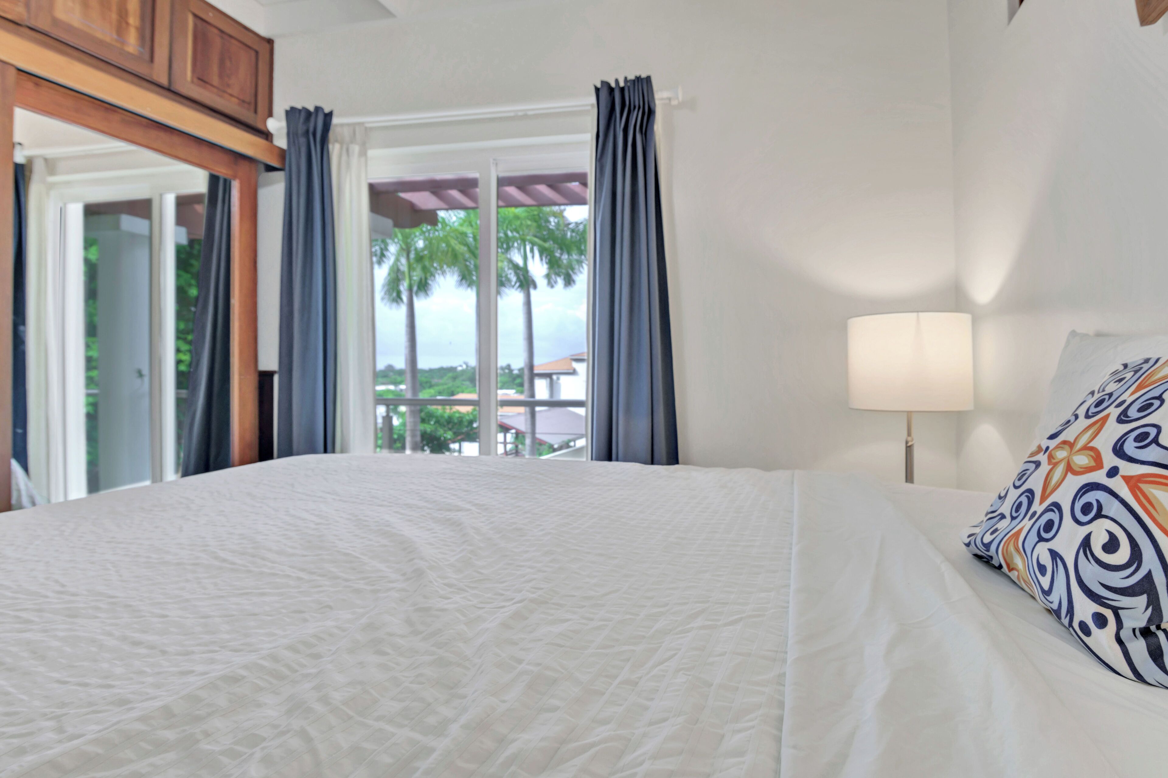 A hotel room with a large bed, a window with curtains, a view of palm trees, and a lamp on the bedside table.