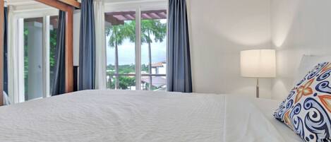 A hotel room with a large bed, a window with curtains, a view of palm trees, and a lamp on the bedside table.