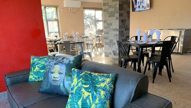 A modern living area with a dark leather sofa, two patterned cushions, a dining table with chairs, and a kitchen area in the background. - The Hill Hotel and Conference Centre (Thohoyandou)