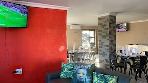 A living room with a red wall, a grey sofa with patterned cushions, a TV mounted on the wall, and a dining area with a table and chairs. - The Hill Hotel and Conference Centre (Thohoyandou)