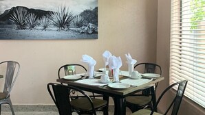 A dining area with a table set for four, chairs, a painting on the wall, and a window with blinds. - The Hill Hotel and Conference Centre (Thohoyandou)