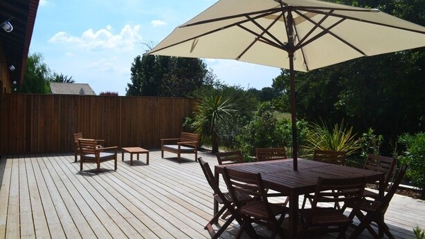 Un patio al aire libre con piso de madera, una mesa redonda con sillas y un gran paraguas que da sombra.
