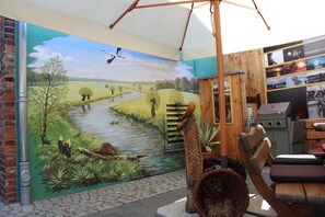 Una habitación rústica con un mural de un paisaje fluvial, una silla de madera y una parrilla.