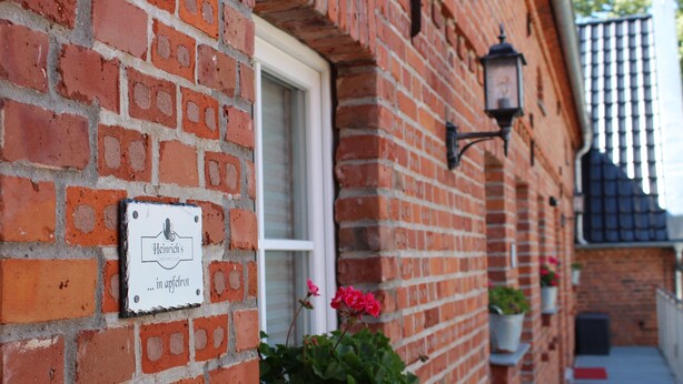 Un bâtiment en briques avec une porte blanche, une enseigne indiquant « Heinrich's in Appeltrot », et des plantes en pot disposées sur le côté.
