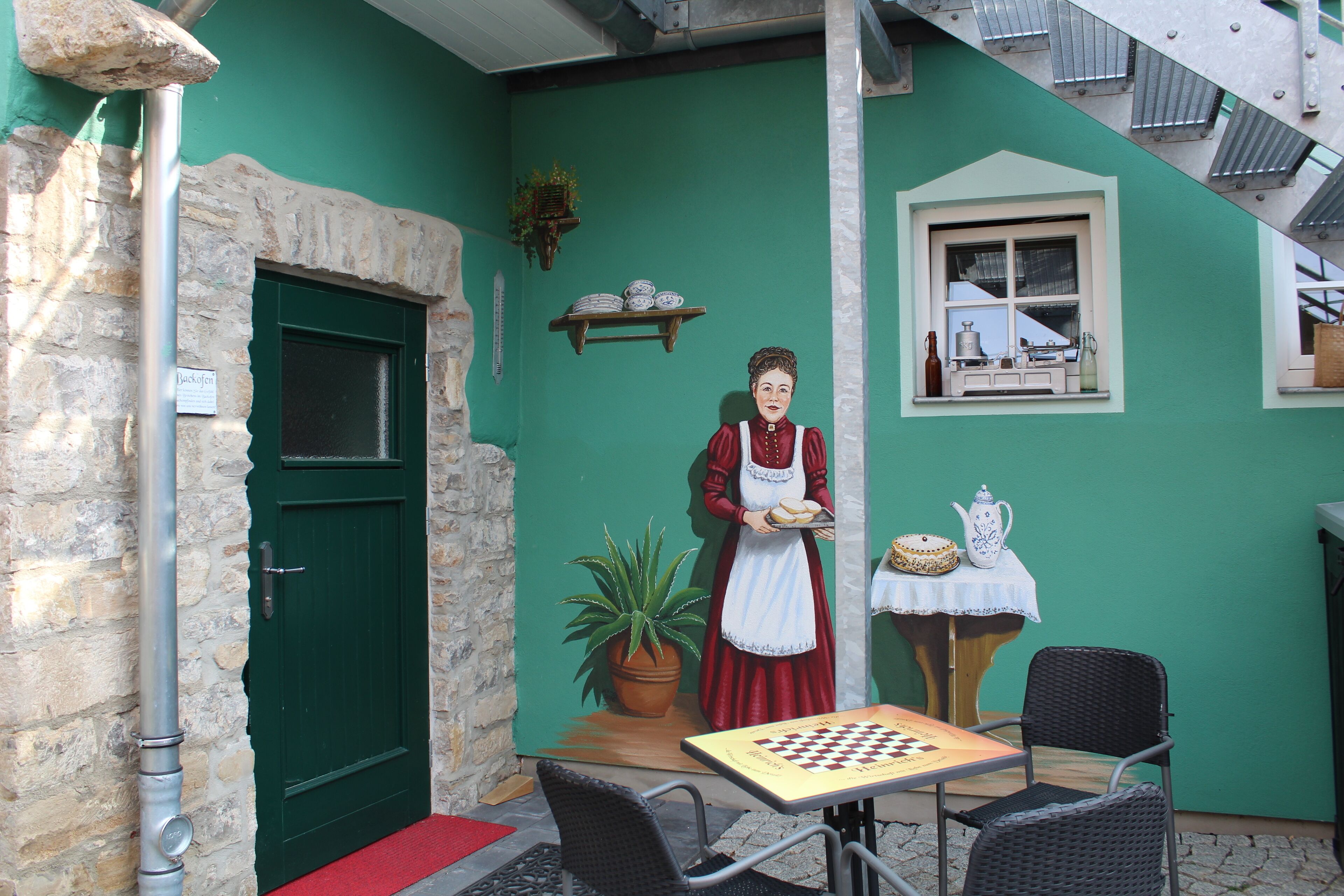 A rustic building with a green door, a stone wall, a mural of a woman holding a tray, a table with a chessboard, and wicker chairs.