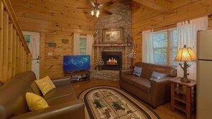 A cozy cabin interior with a stone fireplace, wooden walls, a ceiling fan, and a television. - Cozy cabin, perfect for a honeymoon or an adventure-filled family getaway! (Gatlinburg)