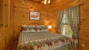 A wooden cabin bedroom with a bed featuring a checkered blanket and moose-themed pillows, a framed map on the wall, and a window with checkered curtains. - Cozy cabin, perfect for a honeymoon or an adventure-filled family getaway! (Gatlinburg)