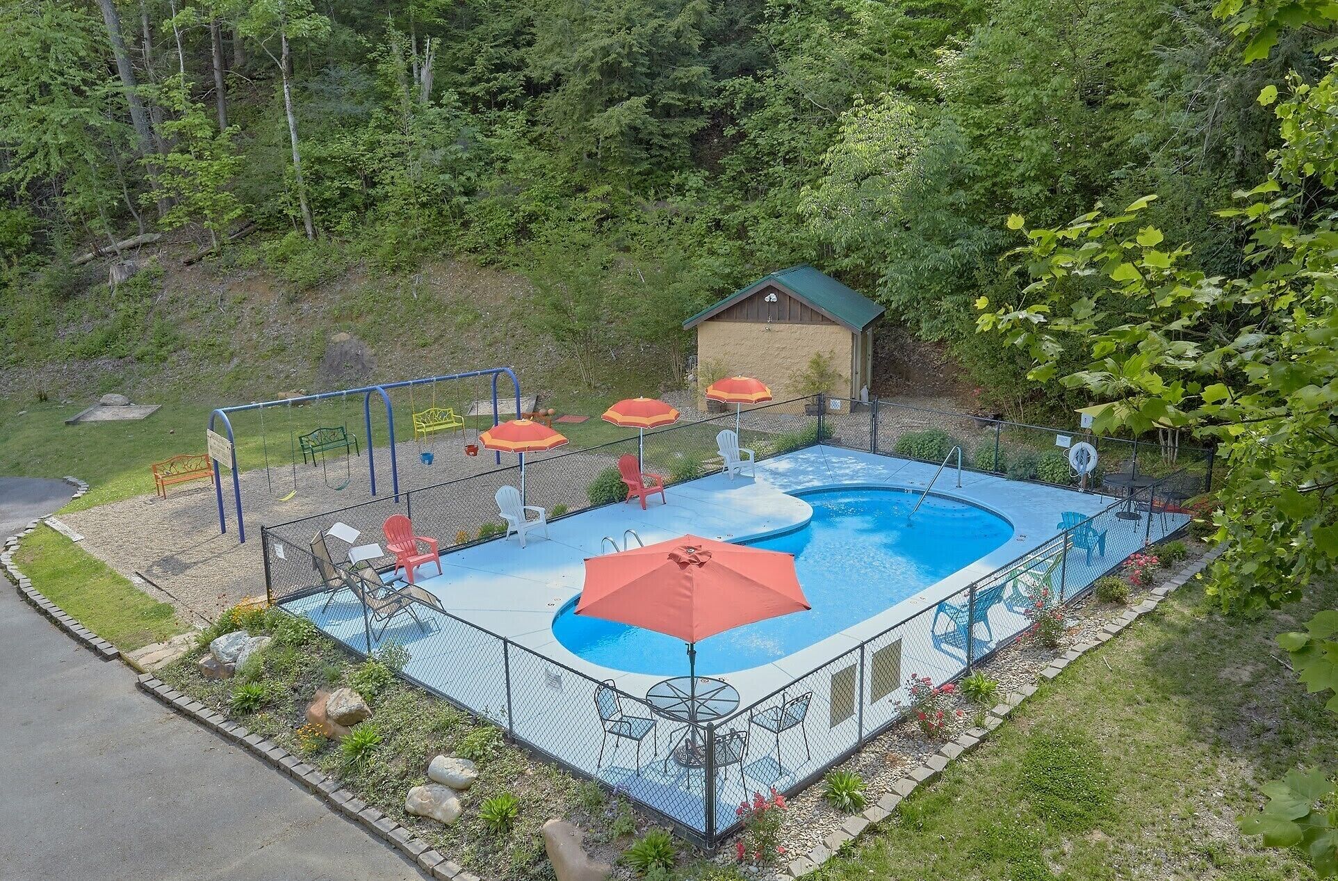A swimming pool with a fenced deck, surrounded by greenery and a small building.
