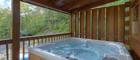 A hot tub in a wooden deck with a view of a swimming pool and trees.