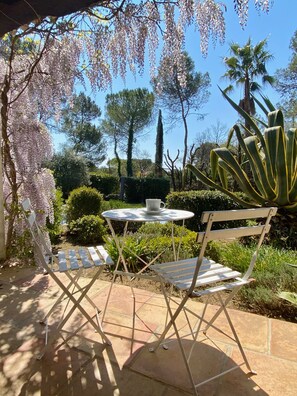 Um pátio com mesa e cadeiras de metal branco, rodeado por vegetação exuberante e diversas plantas.