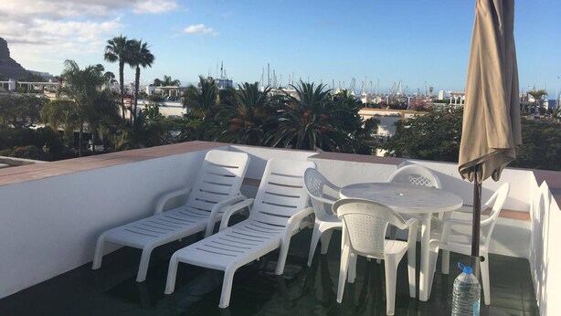 Une terrasse sur le toit, avec des chaises longues en plastique blanc et une table, une bouteille d’eau et un parasol.