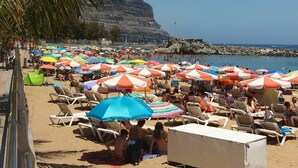 Une plage bordée de rangées de chaises longues et de parasols, un palmier et, au loin, une falaise rocheuse.