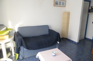 Een kamer met een blauwe bank, een klein tafeltje met een geruit tafelkleed, een gele stoel en een lijst met een afbeelding aan de muur.