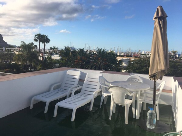 Eine Dachterrasse mit weißen Kunststoff-Lounge-Stühlen und einem Tisch, einer Wasserflasche und einem Sonnenschirm.