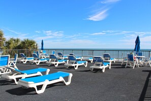 Un área de descanso frente a la playa con filas de tumbonas y sombrillas azules y blancas, una barandilla y vista al mar.