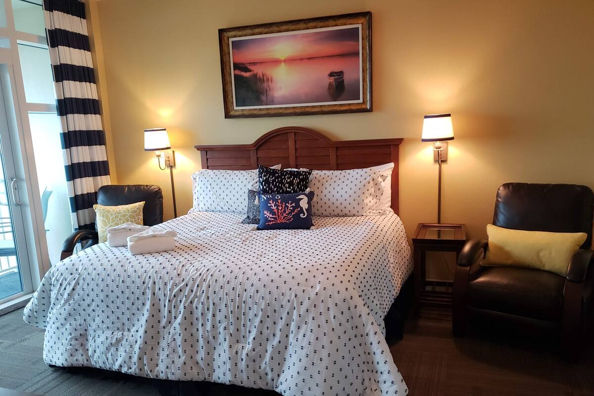 A hotel room with a large bed, two bedside lamps, a wooden headboard, and a painting of a sunset.
