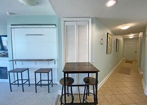 Una cocina con un refrigerador blanco, una mesa de madera con dos taburetes y un ventanal con persianas.
