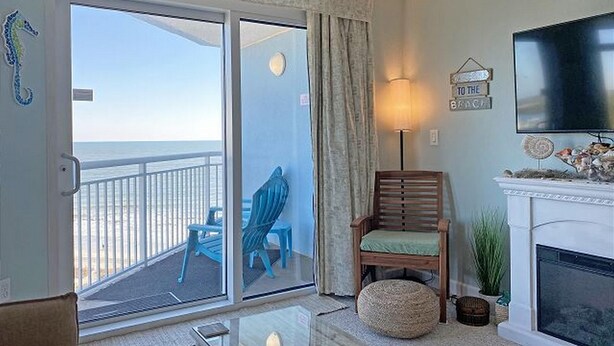 A room with a sliding glass door leading to a balcony with a view of the ocean. There is a wooden chair with a cushion, a floor lamp, and a flat-screen TV mounted on the wall. - Holiday Sands South Resort by Palmetto Vacations (Myrtle Beach)