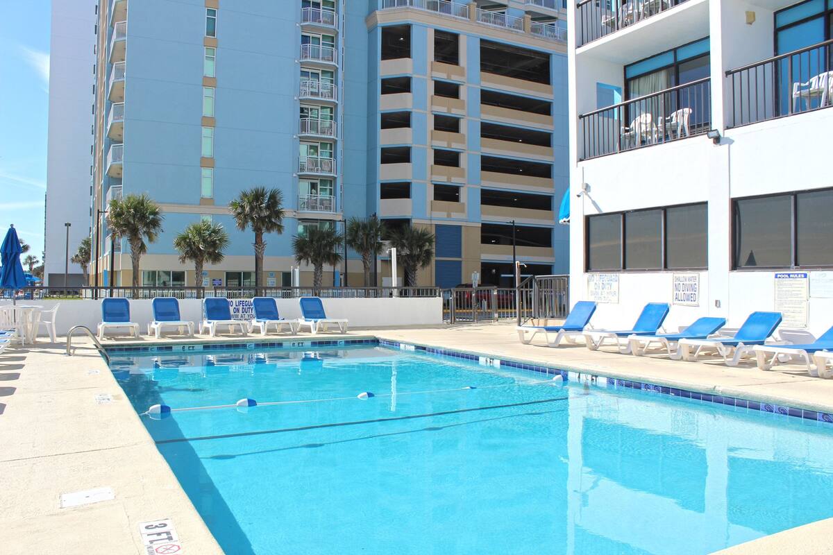 A swimming pool with lounge chairs and palm trees, surrounded by multi-story buildings.