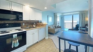 A compact kitchen with white cabinets, a black microwave, and a stove. There is a dining area with a table and chairs, and a balcony with a view. - Holiday Sands South Resort by Palmetto Vacations (Myrtle Beach)