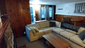 Un salon chaleureux avec un canapé d’angle beige, une table basse en bois et une petite table d’appoint. Un lambris en bois décore l’espace, et une fenêtre avec des rideaux est visible.