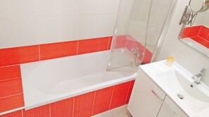 Ein Badezimmer mit einer weißen Badewanne, roten Fliesen, einer Duschkabine aus Glas, einem weißen Waschbecken mit Spiegel und einem gelben Seifenspender.