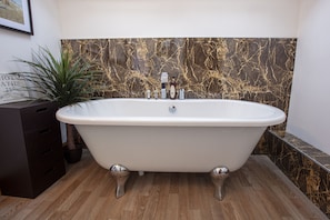 A freestanding bathtub in a bathroom with a marble backsplash and wooden flooring.